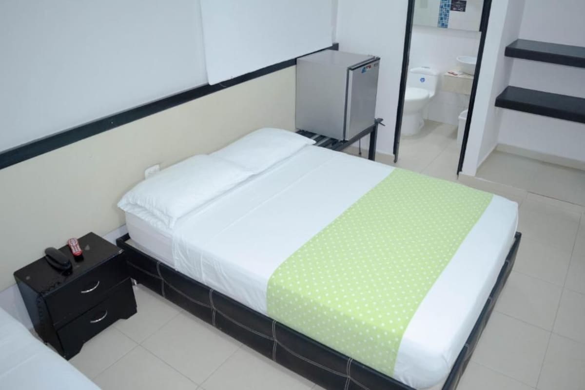 Una cama individual con colchón blanco y manta verde de lunares, una mesita de noche pequeña con un teléfono y un control remoto, y un espejo sobre la cama.