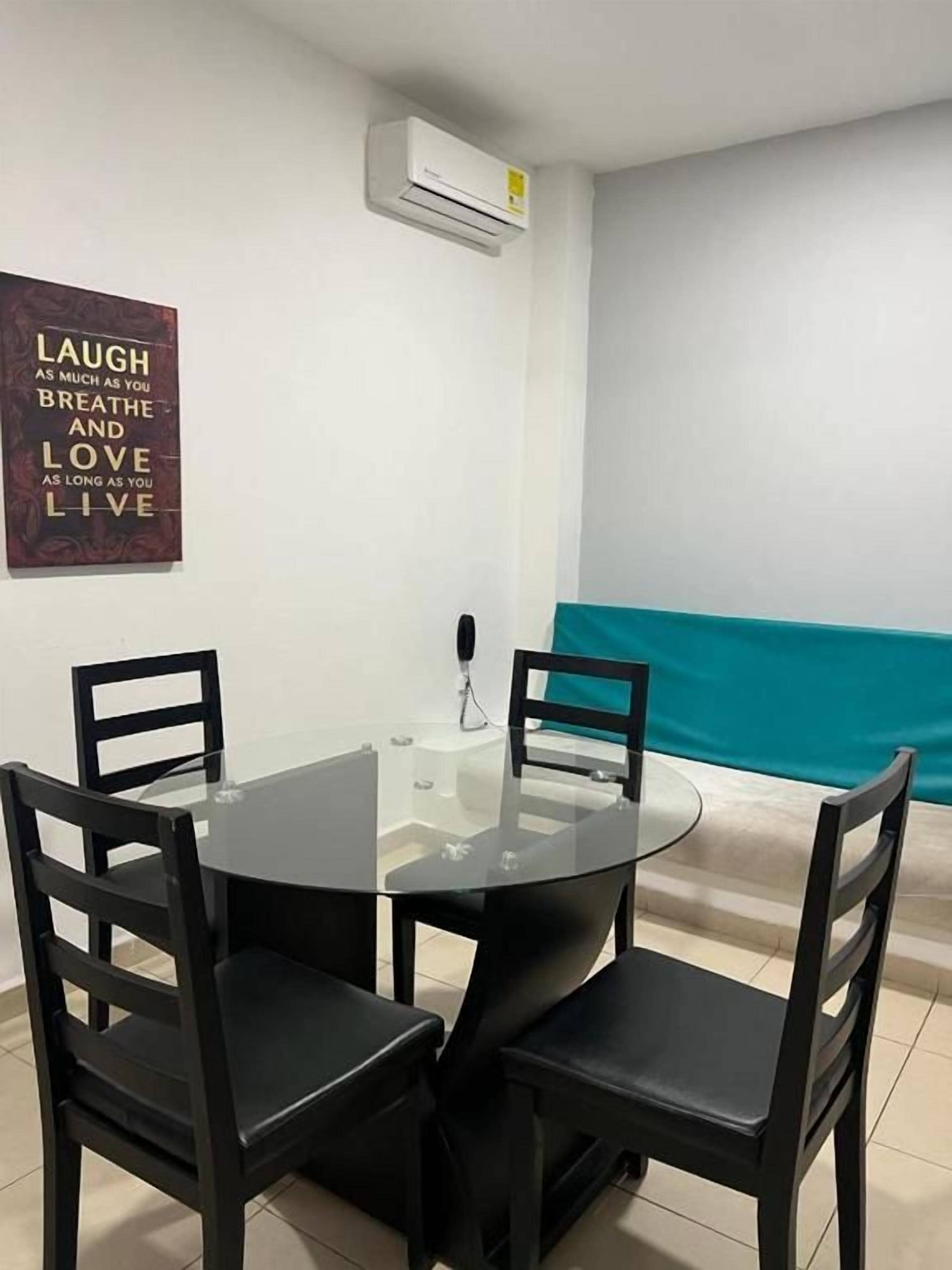 Un comedor con una mesa redonda de vidrio y sillas negras, un aire acondicionado instalado en la pared, y una frase motivadora.
