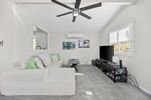 Ein modernes Wohnzimmer mit Deckenventilator, einer Couch mit Kissen, einem Fernseher und Kunstwerken an der Wand.