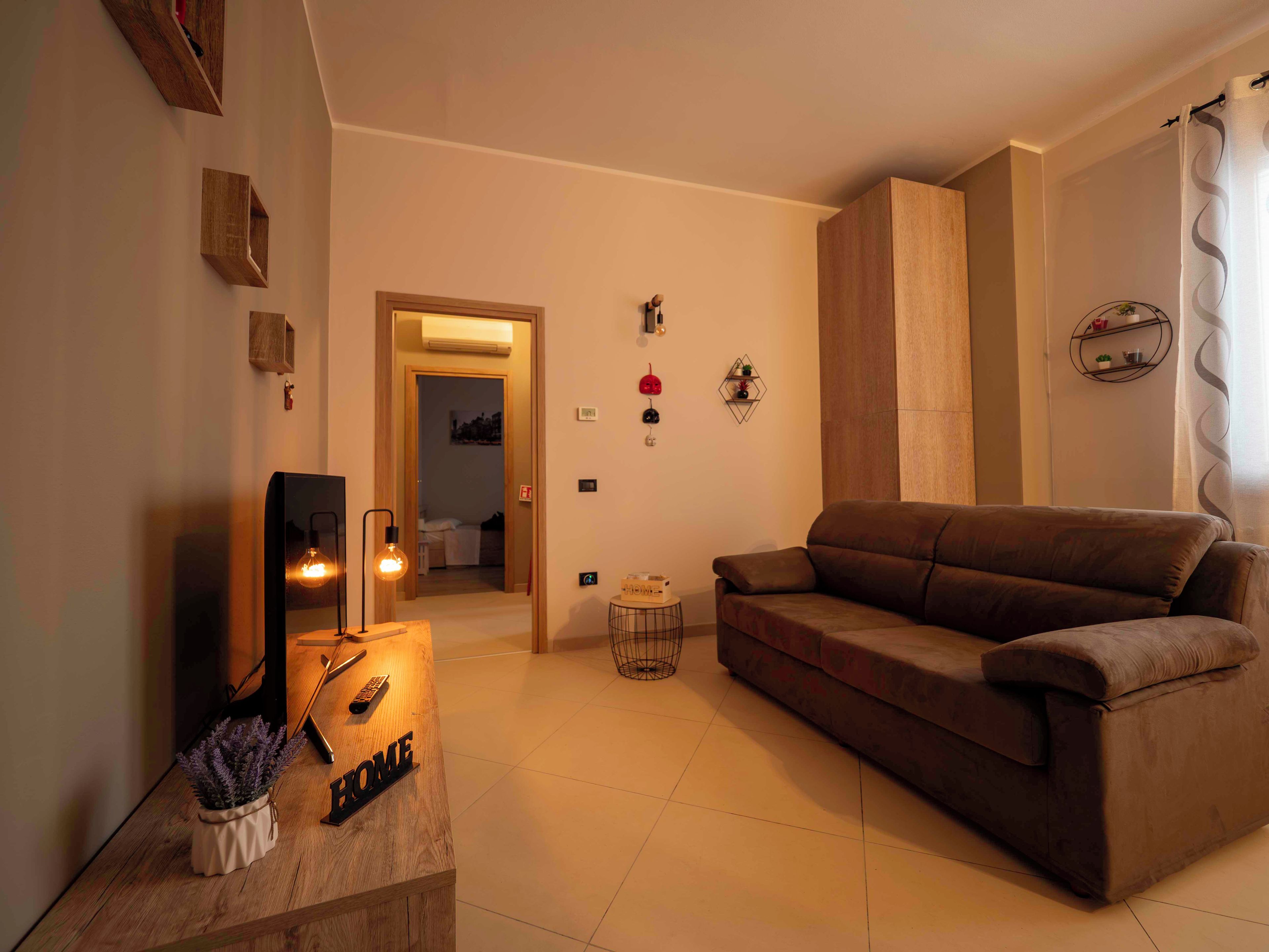 A living room with a sofa, a coffee table with a lamp, and a TV. There is a door leading to another room.