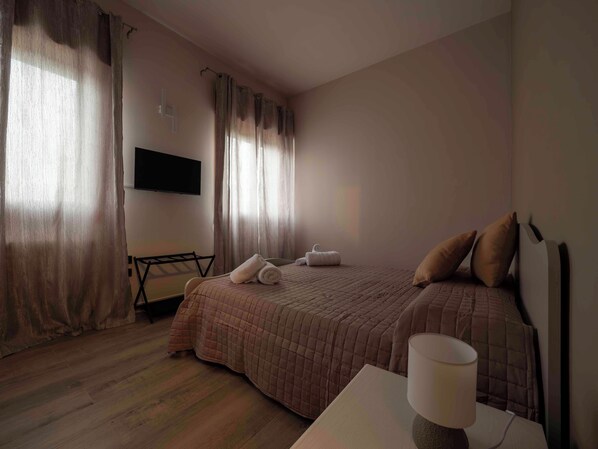 A hotel room with a bed, a TV mounted on the wall, a desk with a lamp, and a window with curtains.