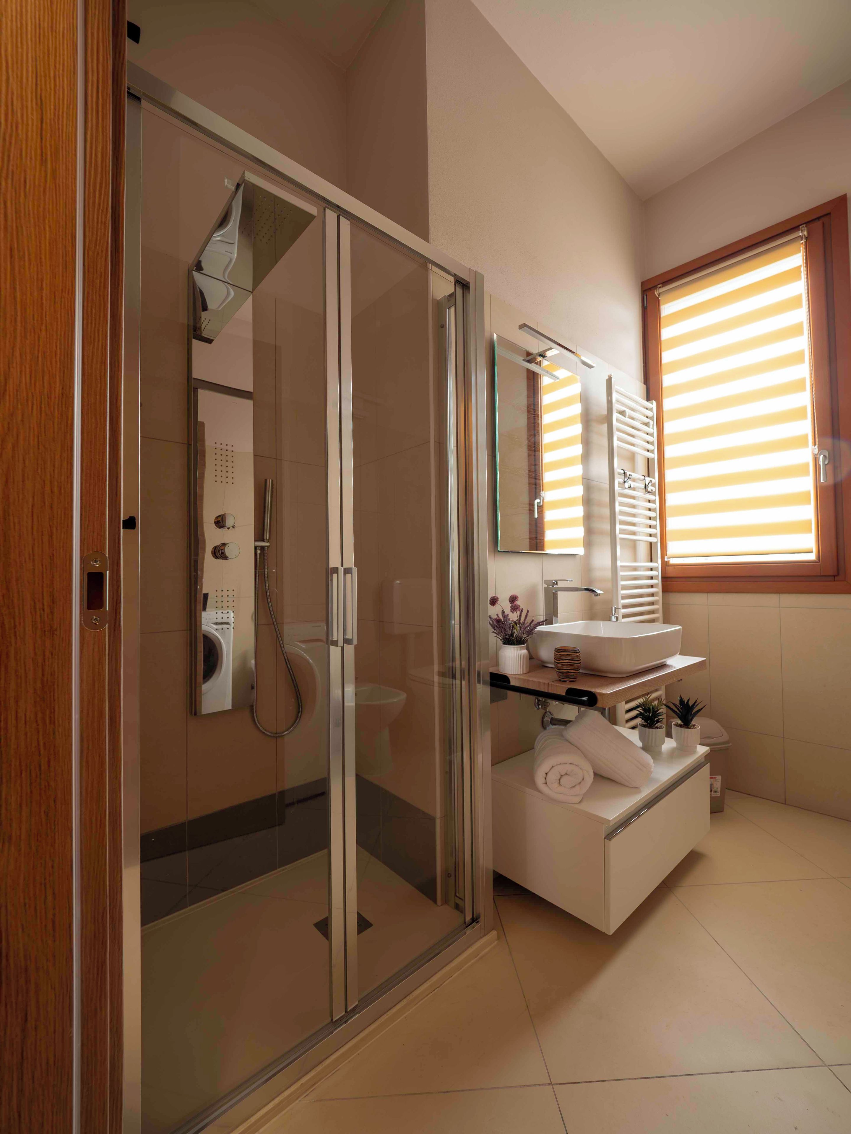 A modern bathroom with a glass-enclosed shower, a white sink, and a window with blinds.
