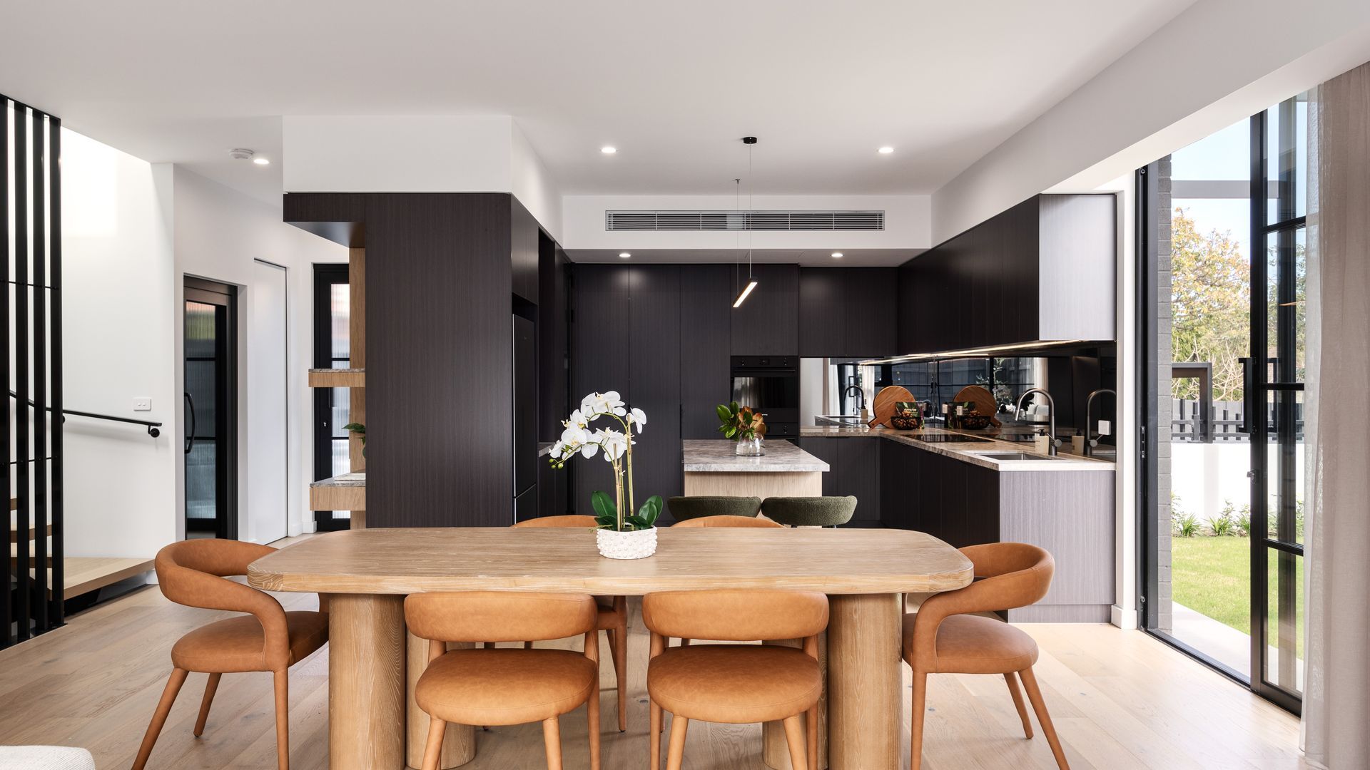 A modern kitchen with a central island, a dining area with a wooden table and chairs, and large windows offering a view of the outdoors.