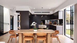 A modern kitchen with a central island, a dining area with a wooden table and chairs, and large windows offering a view of the outdoors. - The Nova - Caulfield South (Caulfield South)