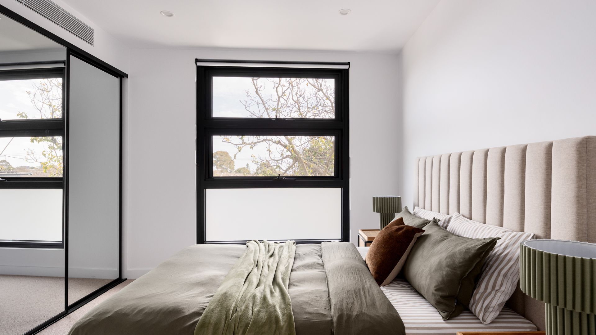 A modern bedroom with a large window, a bed with multiple pillows, and a bedside table with a lamp.
