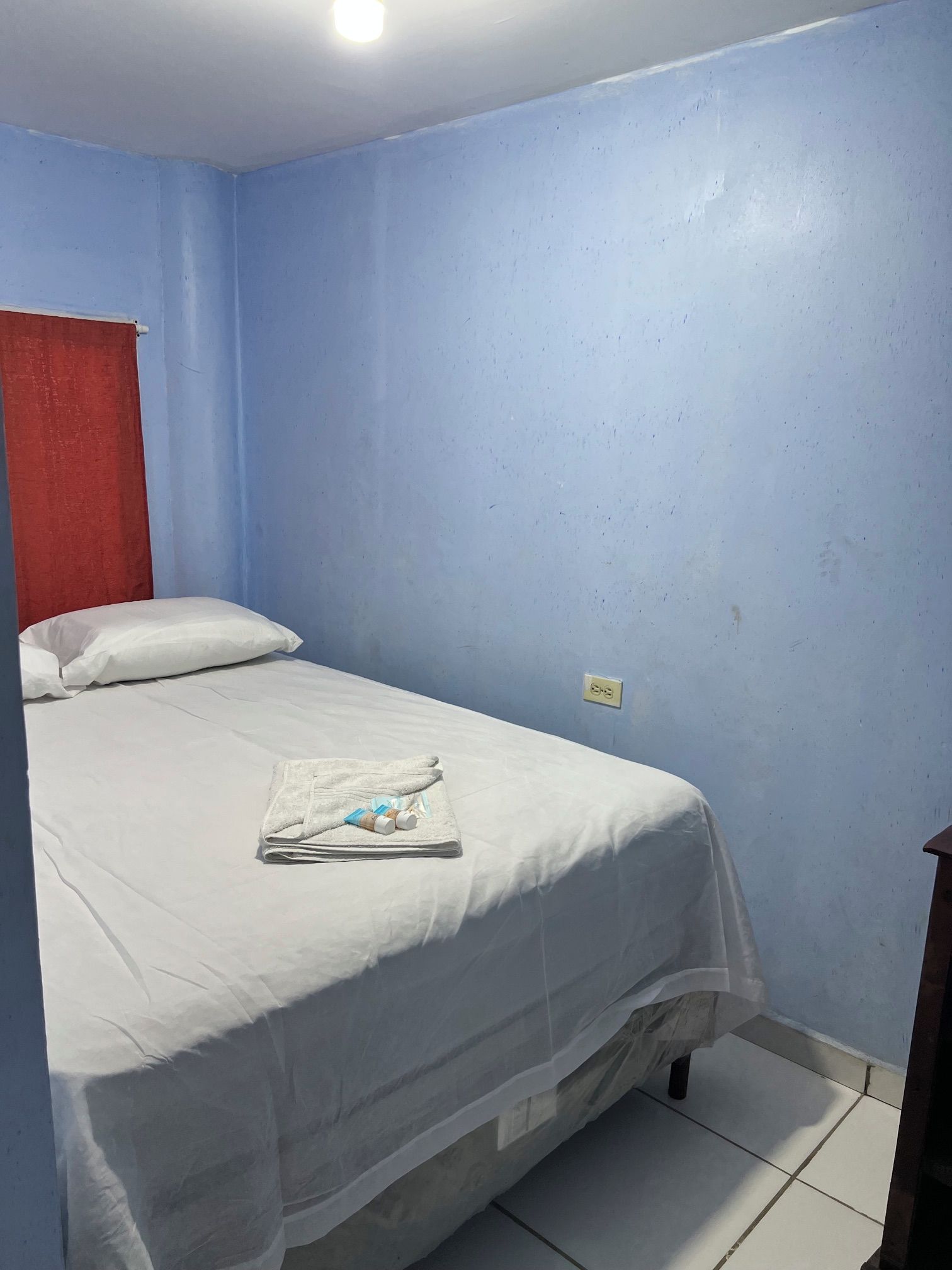 Un lit simple avec une couverture blanche et des oreillers, une serviette pliée et deux bouteilles posées sur le lit.