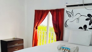 A hotel room with a bed, a nightstand, a window with red curtains, and a wall-mounted air conditioner.
