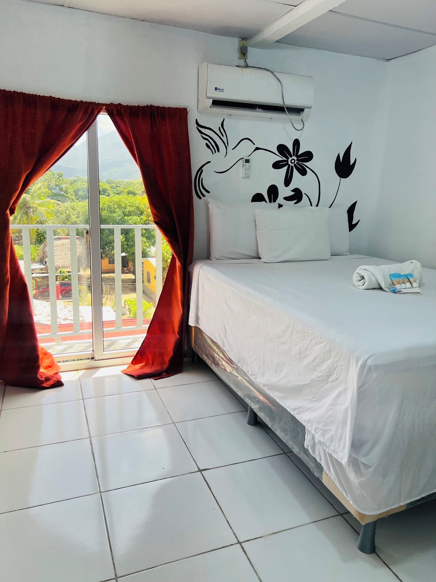 Habitación de hotel con cama, ventana con cortinas rojas, aire acondicionado instalado en la pared y vistas a zonas verdes.