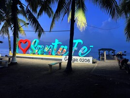 A beachside sign with a heart-shaped cutout, palm trees, a bench, and a paved walkway leading to a pier.