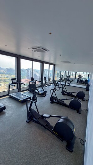A gym with treadmills, elliptical machines, and weight equipment, with large windows offering a city view. - Luxury 3-Bedroom Condo in Tranquil Accra with Pool and Fitness Room (Accra)