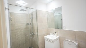 A modern bathroom with a glass shower enclosure, a white sink, and a mirror above it. - Luxury 3-Bedroom Condo in Tranquil Accra with Pool and Fitness Room (Accra)