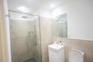 A modern bathroom with a glass shower enclosure, a white sink, and a mirror above it. - Luxury 3-Bedroom Condo in Tranquil Accra with Pool and Fitness Room (Accra)