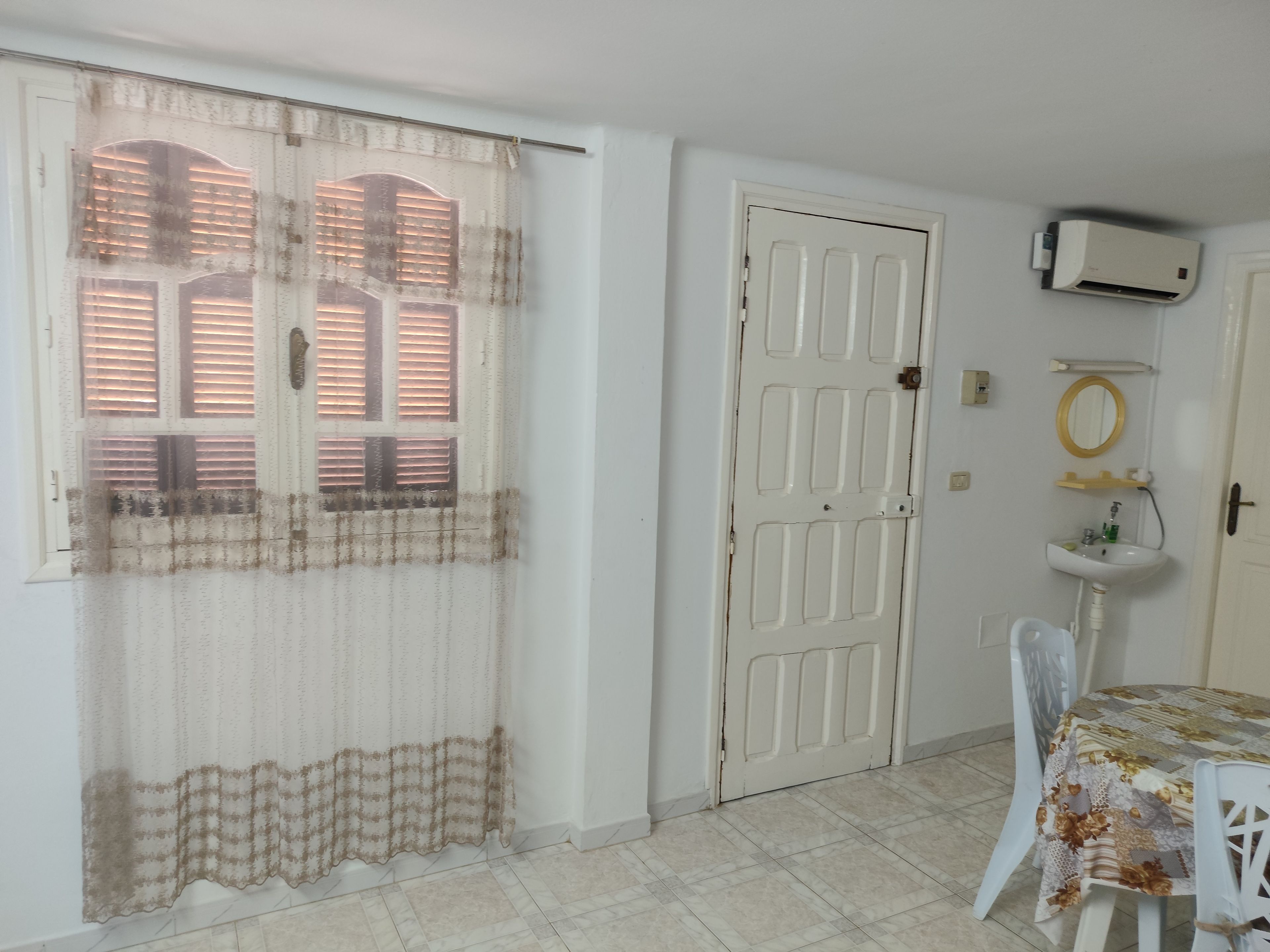 A room with a white door, a window with blinds, a white sink, a mirror, and a table with a floral tablecloth.