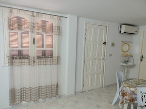 A room with a white door, a window with blinds, a white sink, a mirror, and a table with a floral tablecloth. - Studio Amine  (Mahdia)