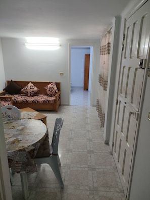 A room with a sofa, a table with chairs, a door, and a hallway leading to another room.