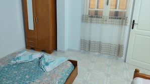 A bedroom with a wooden wardrobe, a bed with floral bedding, a window with curtains, and a white door. - Studio Amine  (Mahdia)