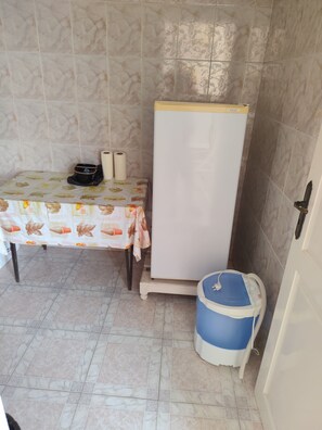 A small kitchenette with a refrigerator, a table with a floral tablecloth, and a blue trash bin. - Studio Amine  (Mahdia)