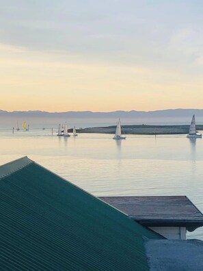 A calm body of water with sailboats, a distant mountain range, and a green-roofed structure in the foreground.