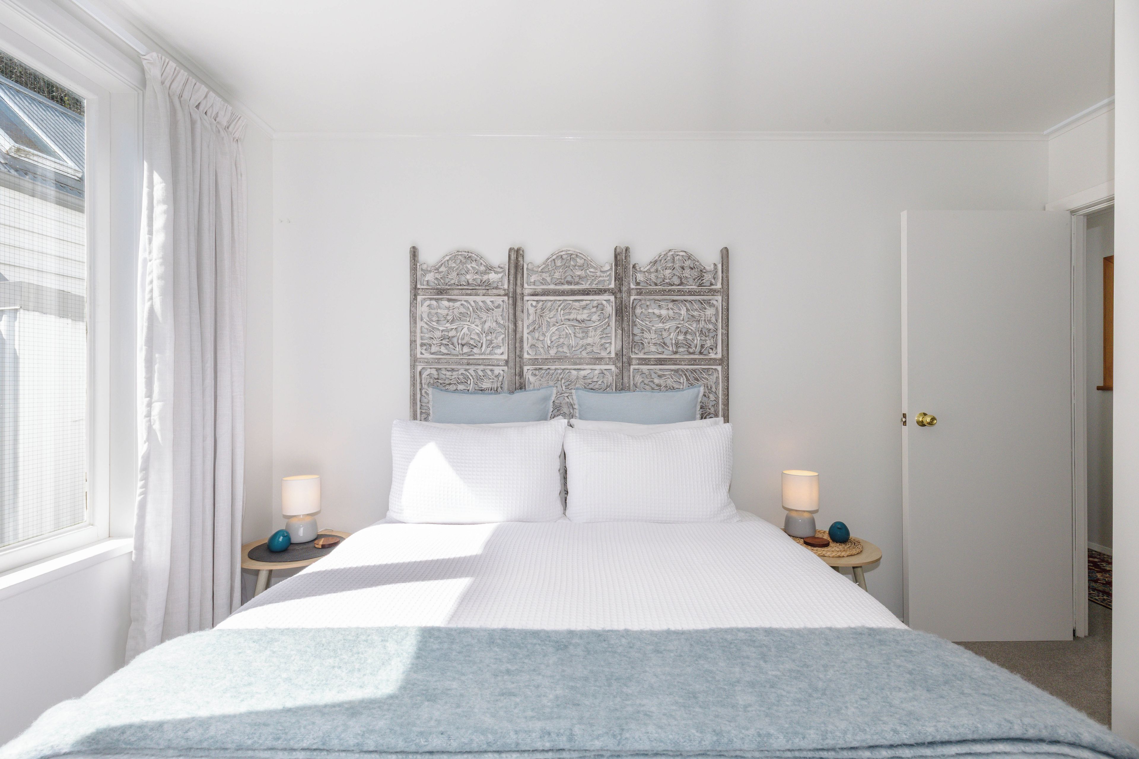 A bedroom with a large bed, two bedside tables, a headboard with intricate carvings, and a window with curtains.