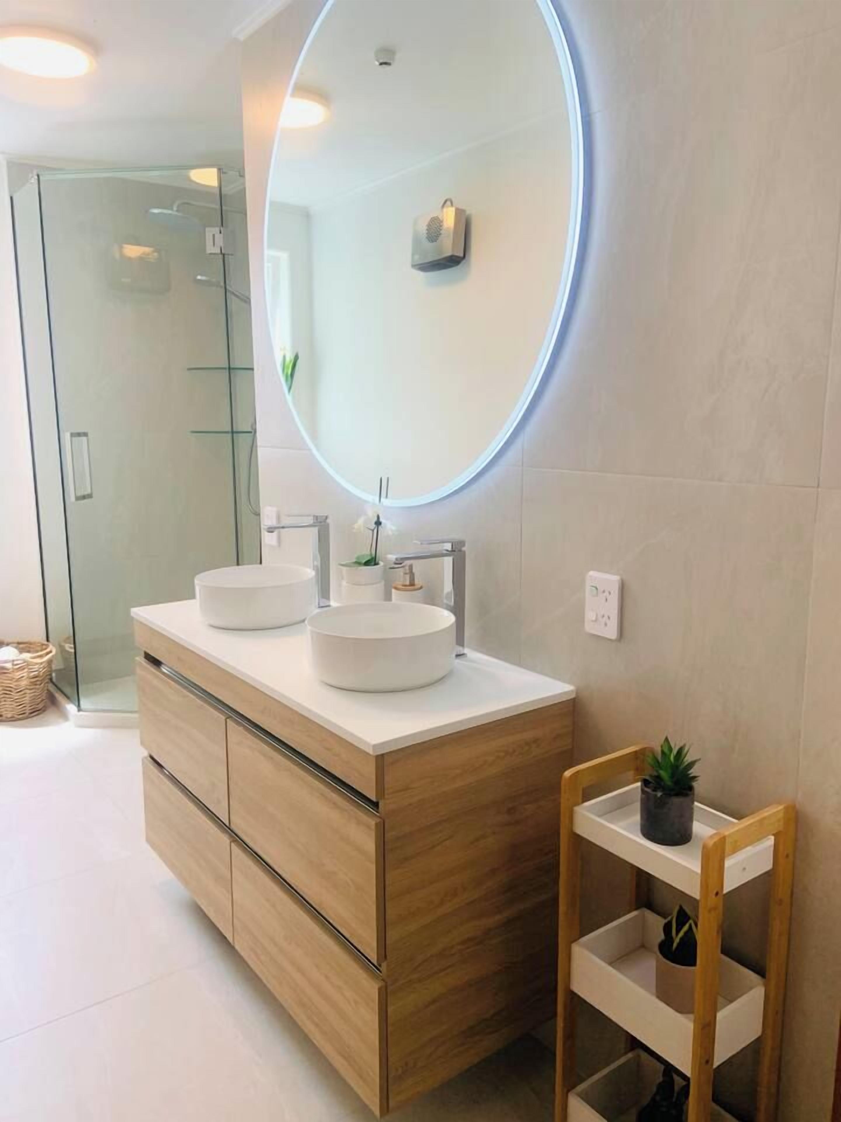 Een moderne badkamer met een houten wastafelmeubel, twee ronde wasbakken, een douchecabine met glazen wanden en een klein houten plankje.
