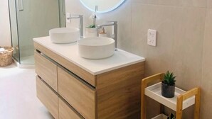 A modern bathroom with a wooden vanity, two round sinks, a glass shower enclosure, and a small wooden shelf. - Nelson Waterfront | Stunning Harbour Views | 2BR (Nelson)