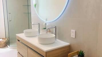 Een moderne badkamer met een houten wastafelmeubel, twee ronde wasbakken, een douchecabine met glazen wanden en een klein houten plankje.