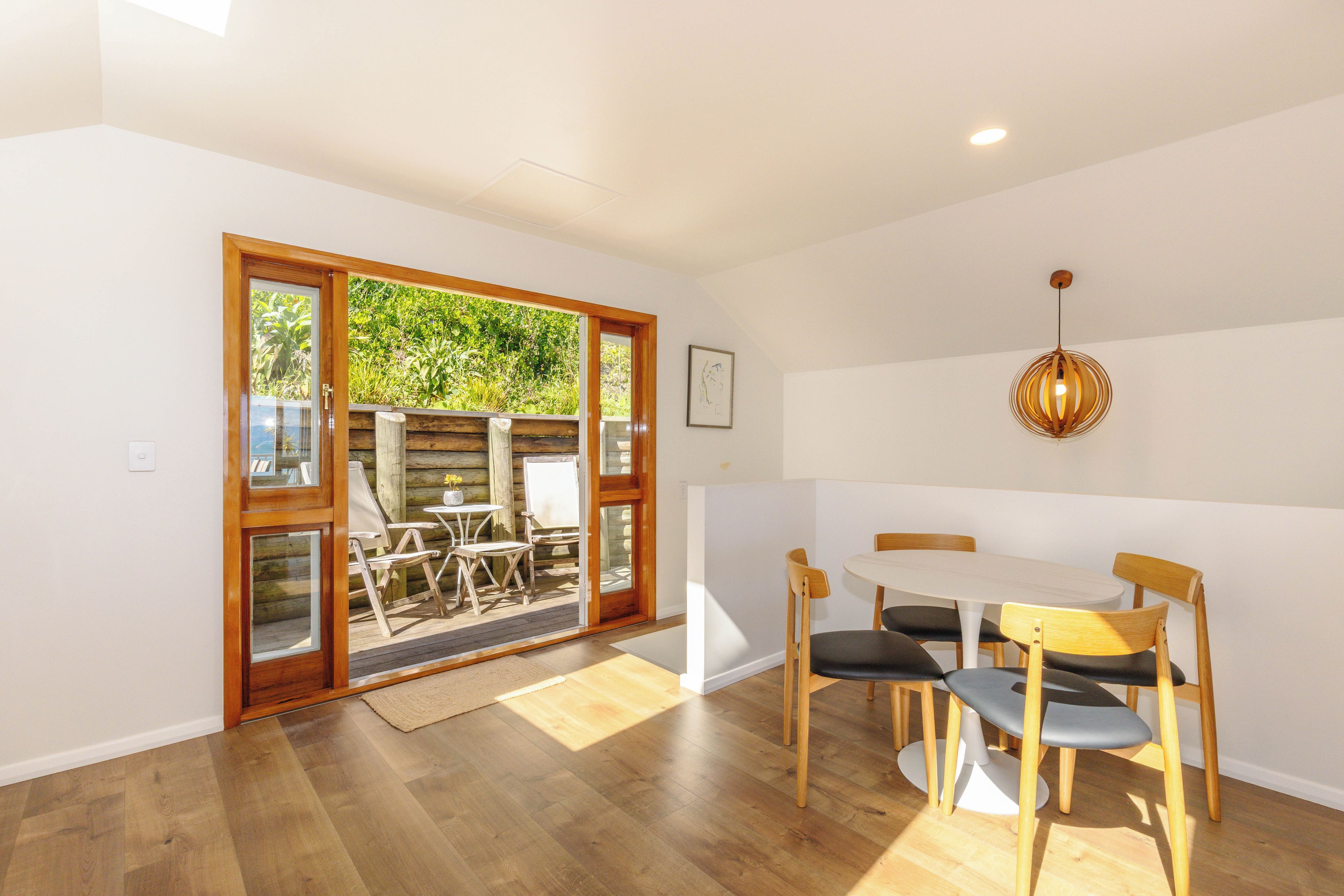 A dining area with a wooden table and chairs, a hanging light fixture, and a view of greenery through a glass door.