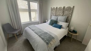 A bedroom with a bed, a chair, a bedside table, and a window with curtains. - Nelson Waterfront | Stunning Harbour Views | 2BR (Nelson)