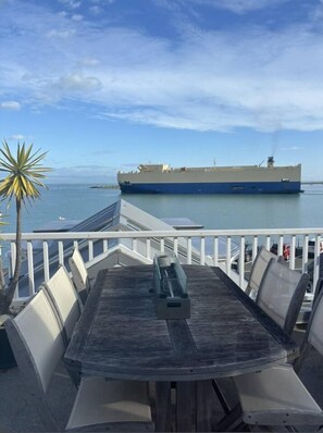 Ein Holztisch mit Stühlen, einer Geländer und ein Blick auf ein großes Schiff in der Ferne.