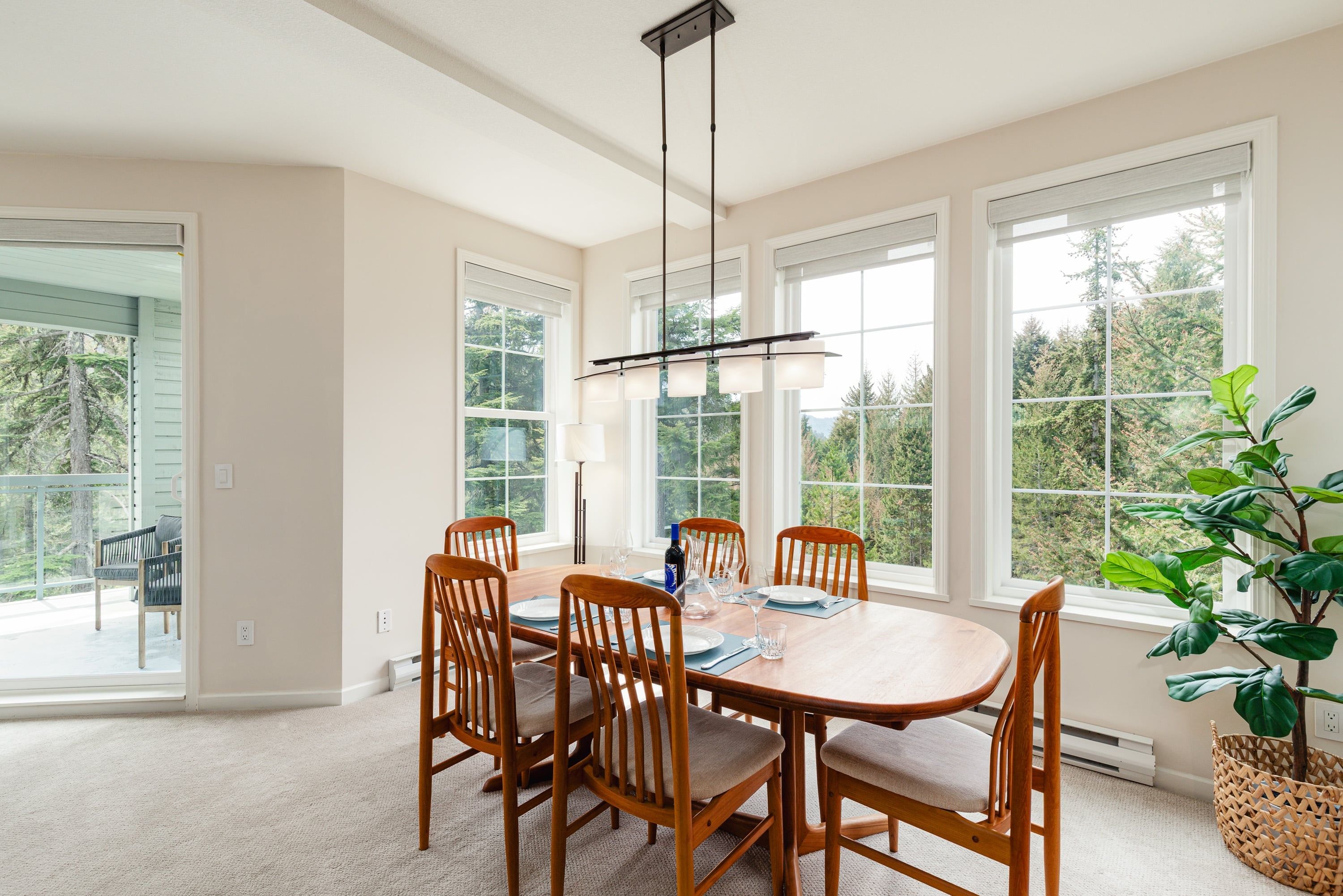 A dining area with a wooden table and chairs, large windows, and a hanging light fixture.