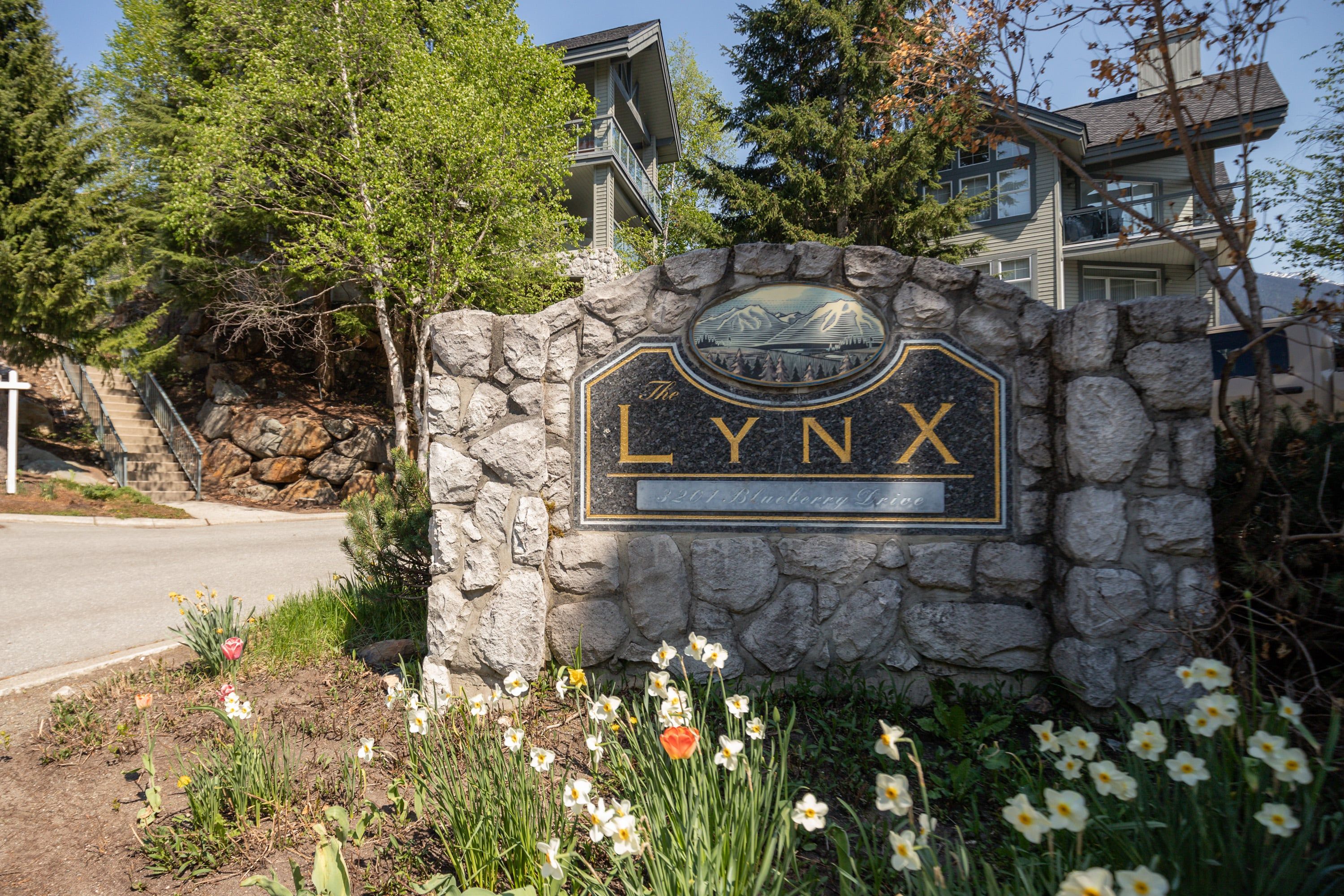 Ein Steinschild mit der Aufschrift 'LYNX' und einer Berglandschaft, umgeben von Blumen und Bäumen.