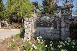 Un letrero de piedra con la palabra 'LYNX' y un paisaje montañoso, rodeado de flores y árboles.
