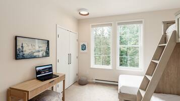 A bedroom with a bed, a desk with a laptop, a window with a view of trees, and a door.