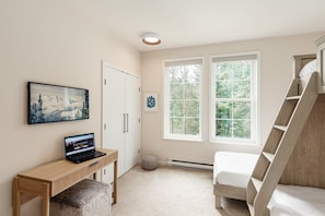Un dormitorio con cama, un escritorio con una computadora portátil, una ventana con vista a árboles y una puerta.