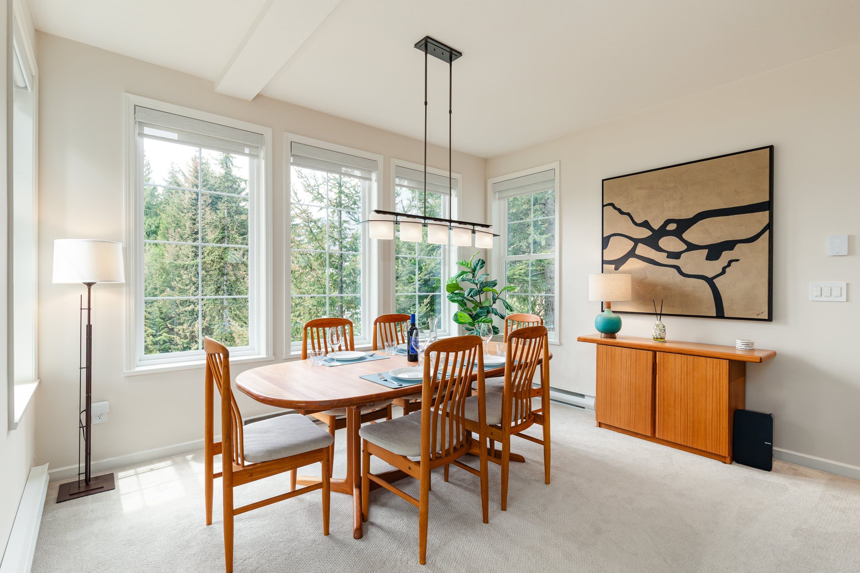 A dining area with a wooden table and chairs, large windows, a hanging light fixture, and a painting on the wall.