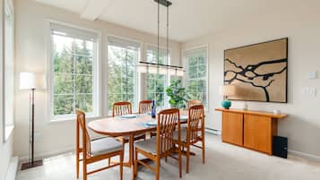 A dining area with a wooden table and chairs, large windows, a hanging light fixture, and a painting on the wall.