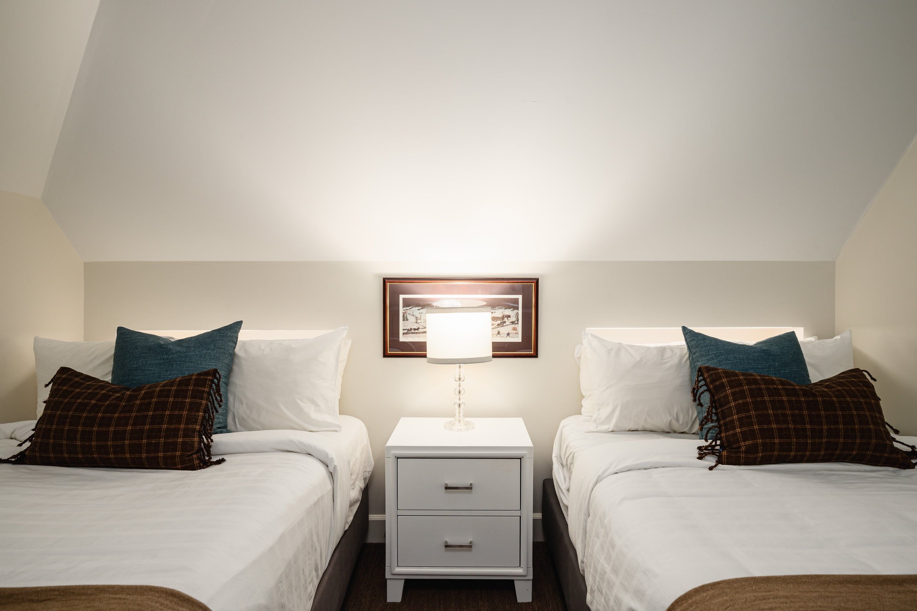A twin room with two beds, a nightstand with drawers, a framed picture above the nightstand, and a lamp.