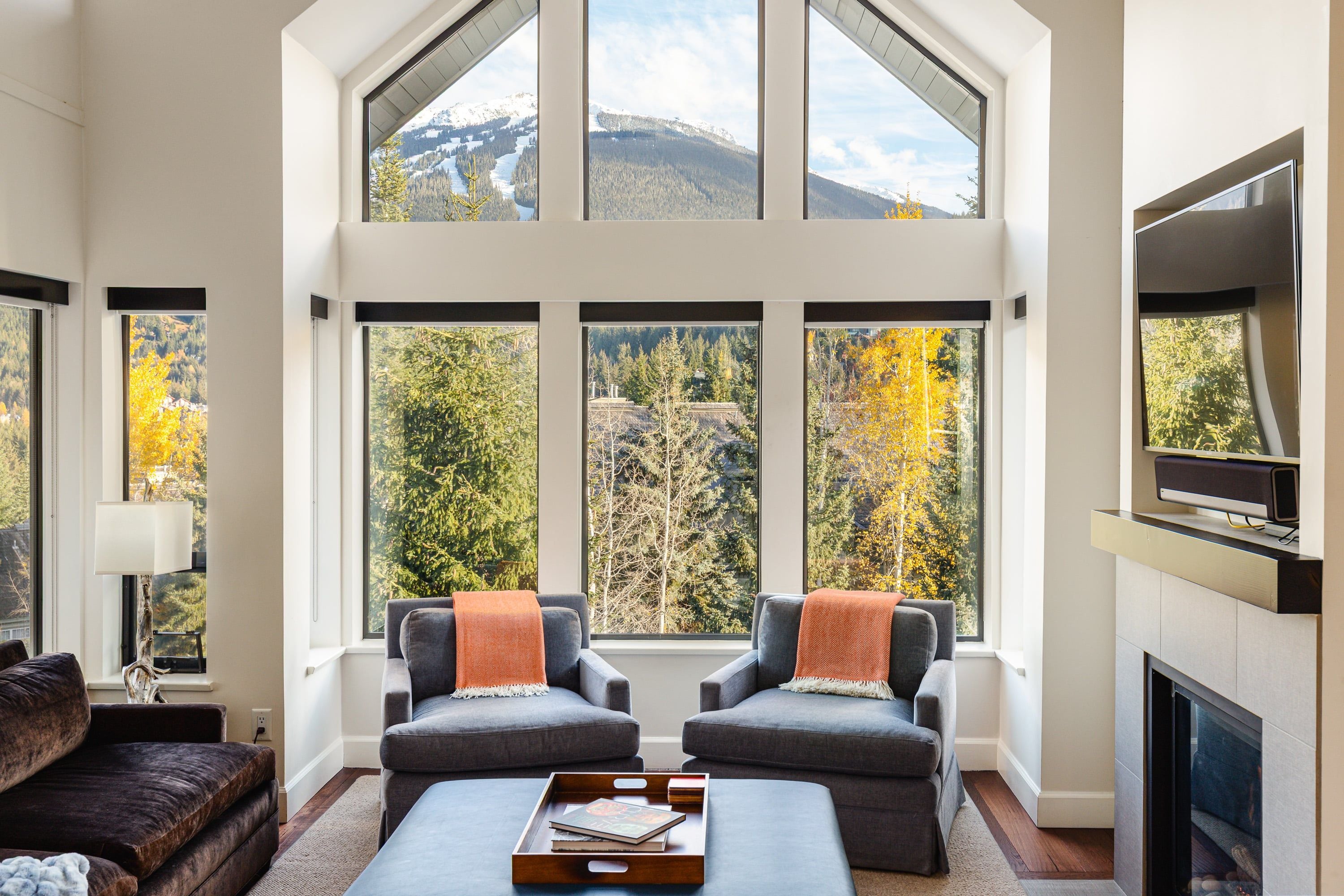 A spacious living room with a high ceiling, large windows offering a view of trees and mountains, two grey armchairs with orange throw pillows, a dark coffee table with a tray, and a flat-screen TV mounted on the wall.