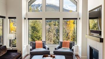 A spacious living room with a high ceiling, large windows offering a view of trees and mountains, two grey armchairs with orange throw pillows, a dark coffee table with a tray, and a flat-screen TV mounted on the wall.