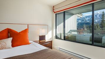 A hotel room with a large window offering a mountain view, a bed with orange pillows, and a bedside table with a lamp.