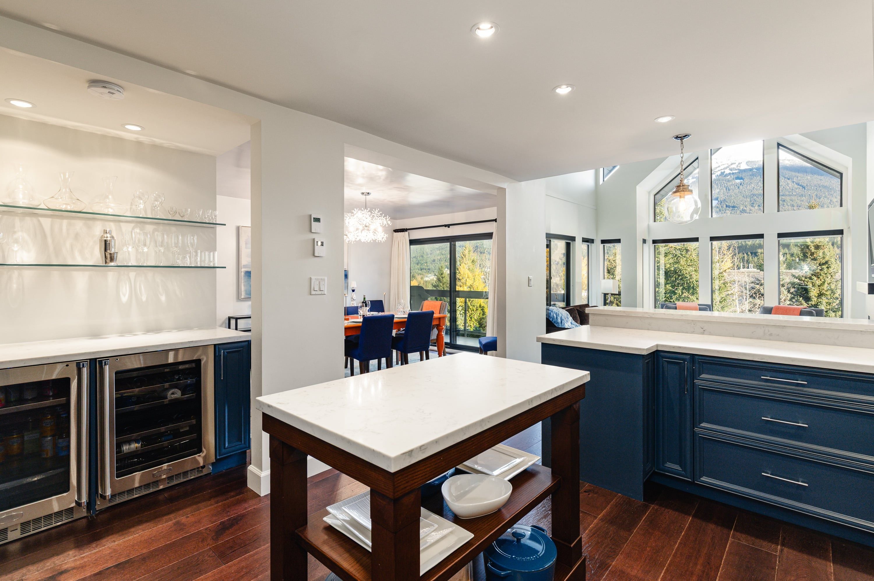 A modern kitchen with dark blue cabinetry, a marble countertop, and a dining area with a view of the outdoors.