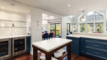 A modern kitchen with dark blue cabinetry, a marble countertop, and a dining area with a view of the outdoors.
