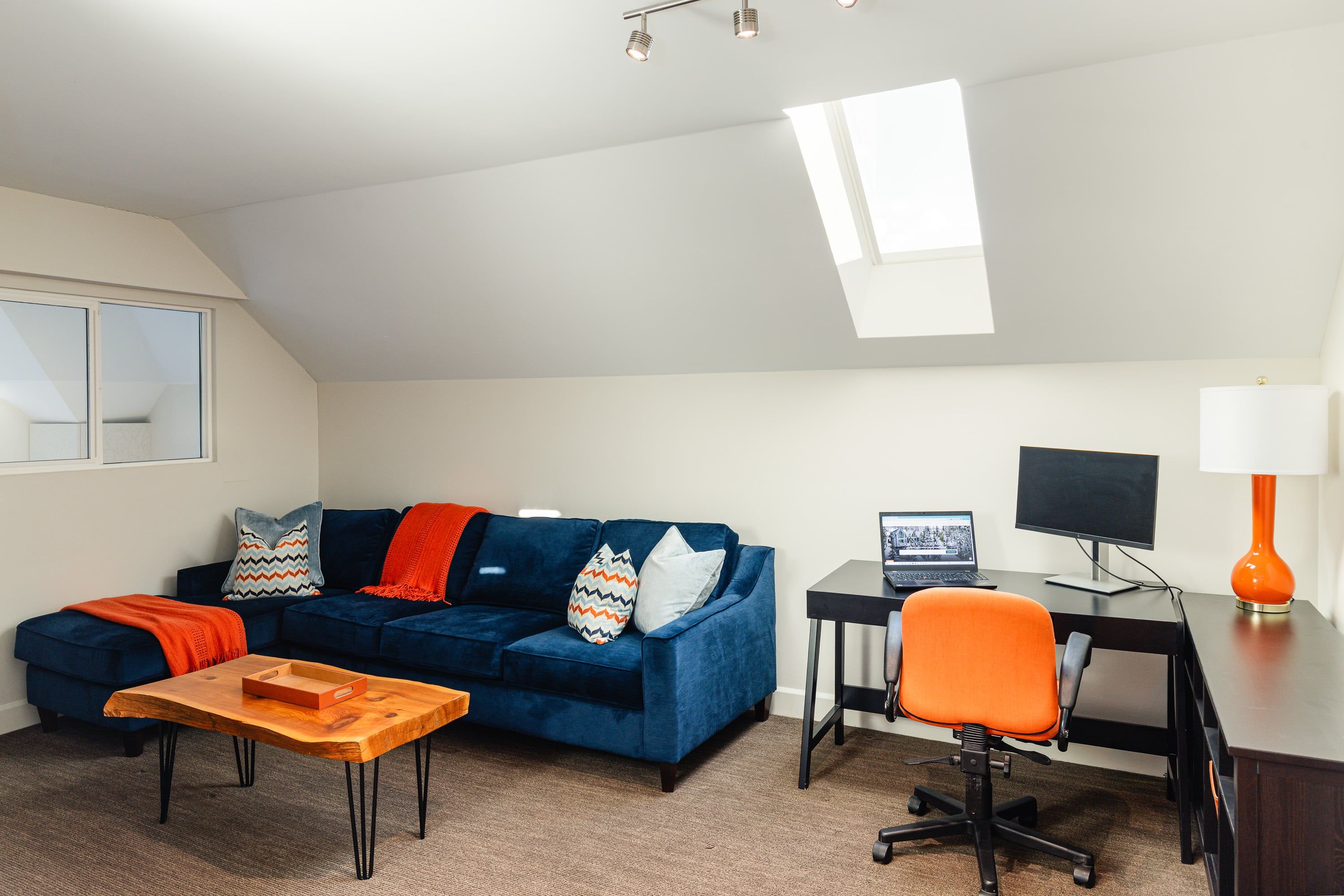 A modern living room with a blue sofa, a wooden coffee table, a desk with a computer, and an orange chair.