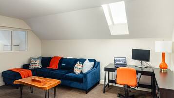 A modern living room with a blue sofa, a wooden coffee table, a desk with a computer, and an orange chair.