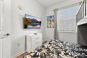 Un dormitorio con una cama, un aparador, un cuadro de un paisaje montañoso, un cuadro de una persona con globos y una ventana con persianas.