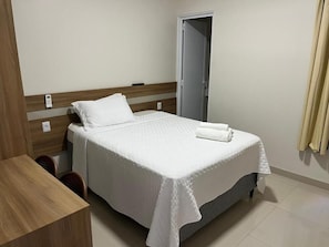 Una cama bien tendida con una colcha blanca y dos almohadas, un cabecero de madera, una mesita de noche y una puerta al fondo.