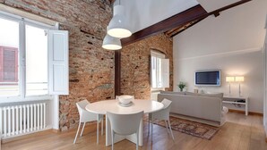 Ein modernes Interieur mit Backsteinwand, einer Essecke mit weißem Tisch und Stühlen, einer Couch, einem Fernseher und einer Lampe.