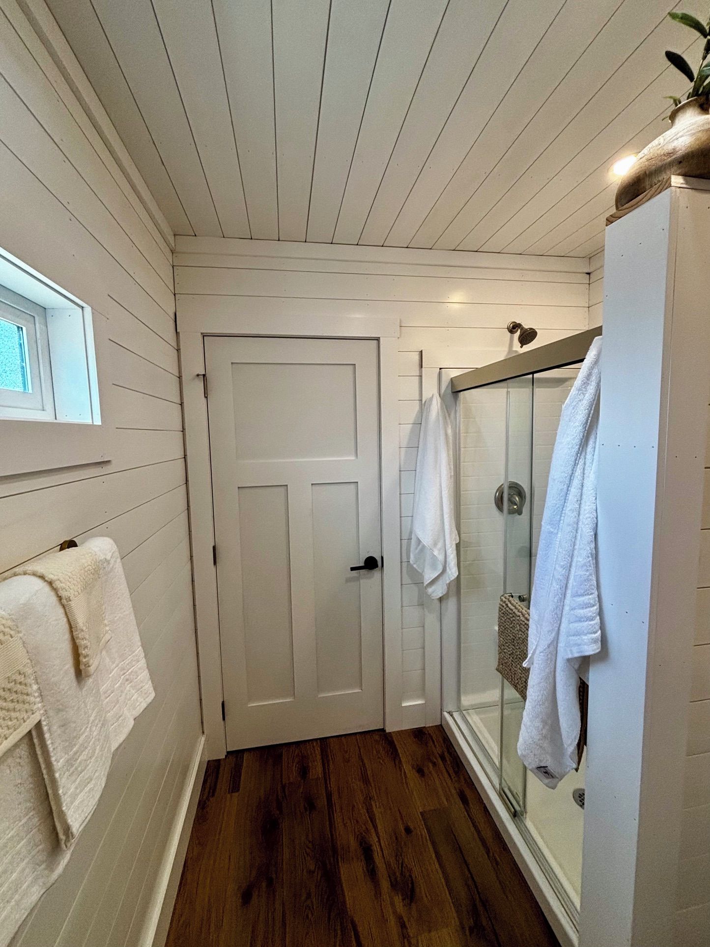 A narrow bathroom with a wooden floor, a white door, and a glass shower enclosure.