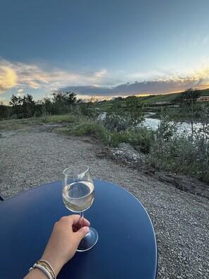 Une main tenant un verre à vin sur une table bleue, avec en arrière-plan une rivière et des collines.