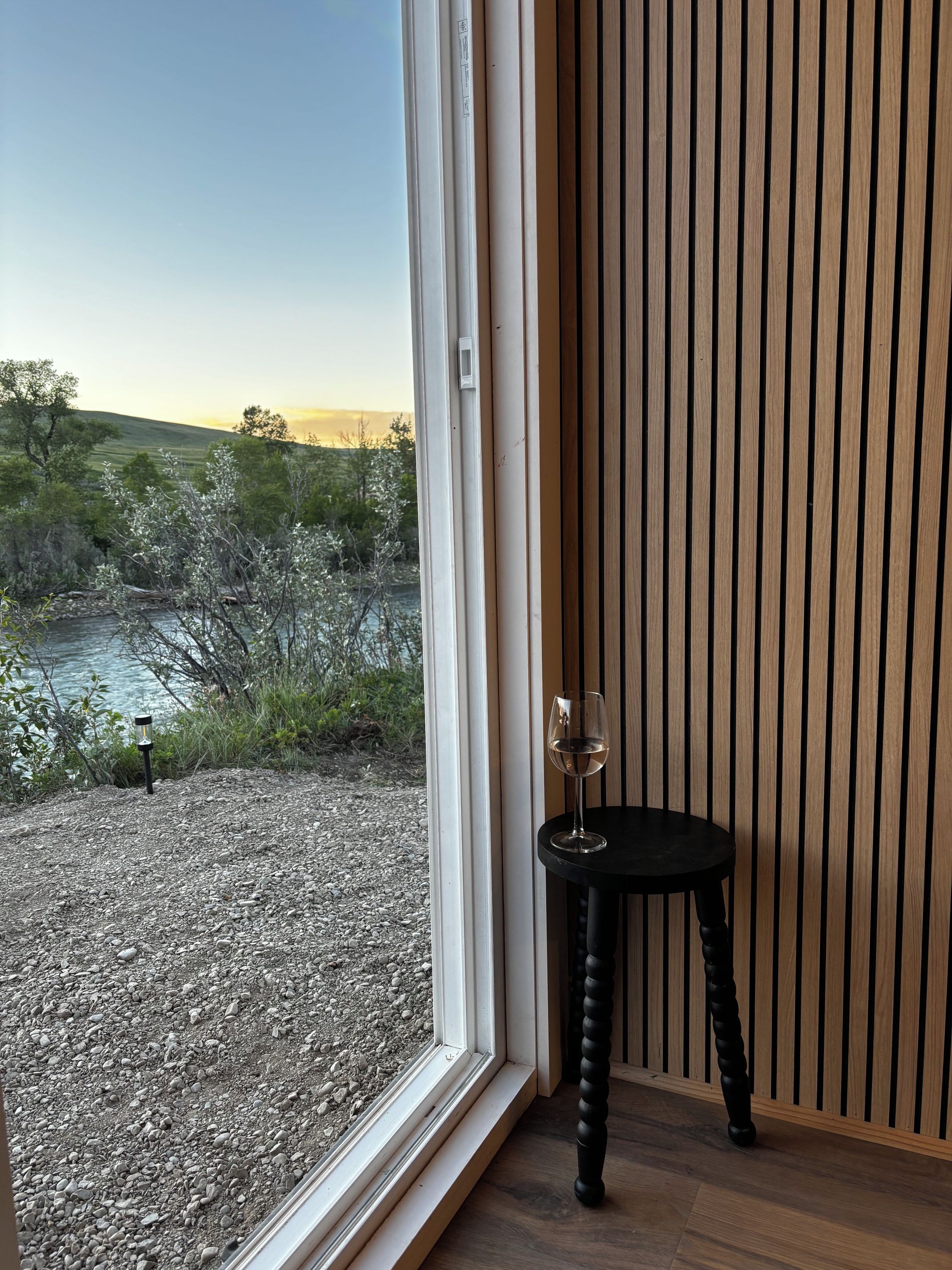 Een kamer met een groot raam dat uitkijkt op een natuurlandschap, een klein tafeltje met een glas wijn, en een wand van houten latten.